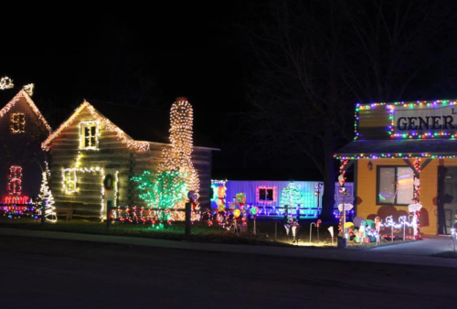 A festive scene of a log cabin and a general store, both adorned with colorful holiday lights at night.
