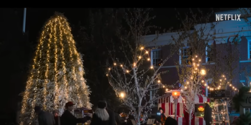 A festive scene with a lit Christmas tree and decorated trees, people gathered around, and a food truck nearby.