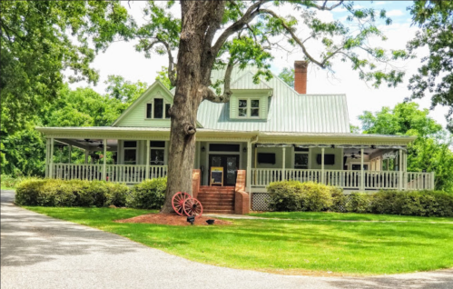 A charming green house with a porch, surrounded by trees and a manicured lawn, featuring a decorative wagon wheel.