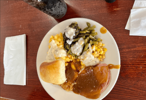 A plate of food featuring corn, green beans with sauce, a biscuit, and meat with gravy on a wooden table.