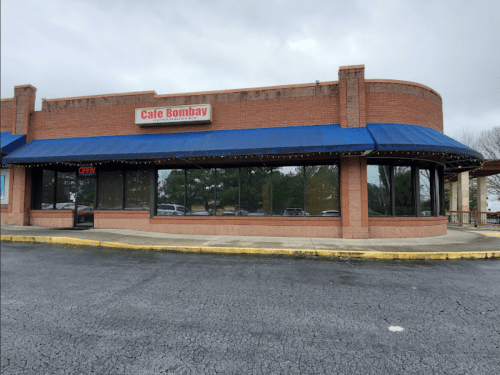 Exterior of Cafe Bombay, featuring a blue awning and "OPEN" sign, set in a shopping plaza on a cloudy day.