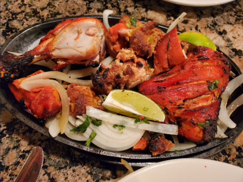 Sizzling platter of grilled chicken pieces with onions, lime wedges, and herbs on a stone surface.