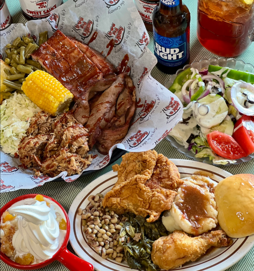 A spread of barbecue dishes including ribs, pulled pork, fried chicken, sides, and drinks on a table.