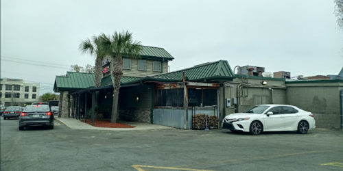 A restaurant with a green roof, palm trees, and parked cars in a lot on a cloudy day.
