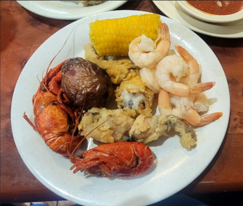 A plate of seafood featuring crawfish, shrimp, fried fish, a corn on the cob, and a stuffed potato.
