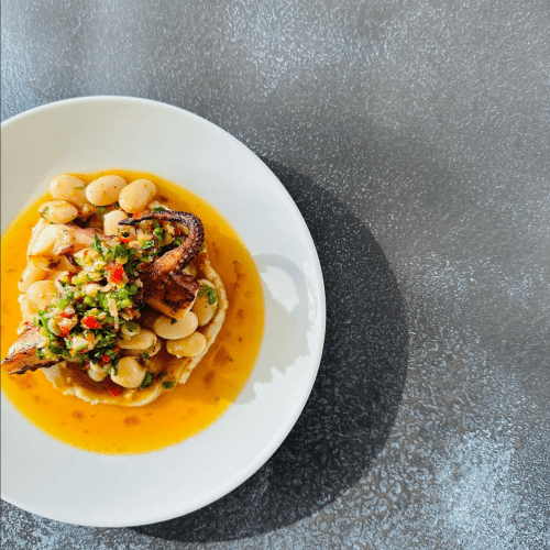 A plate of grilled fish atop a bed of beans, garnished with a colorful salsa, served in a vibrant sauce.
