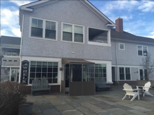 A coastal building with a gray shingle exterior, featuring large windows and a patio with adirondack chairs.