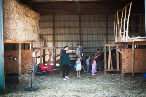 A family gathers in a barn, with hay bales and children playing in a rustic, sunny environment.