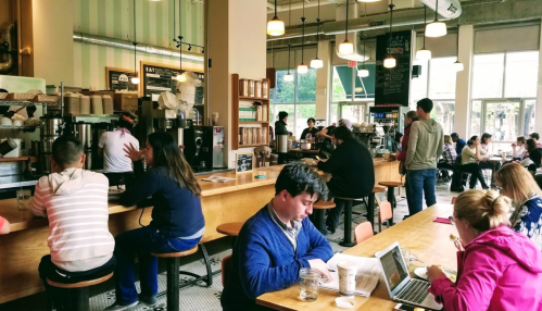 A busy café with people working, chatting, and enjoying drinks at wooden tables and a long counter.