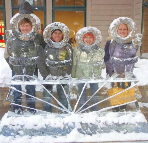 Four people stand behind an ice sculpture of astronauts, surrounded by snow.