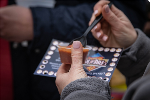 A close-up of a hand holding a small cup of sauce with a black spoon, resting on a promotional card.