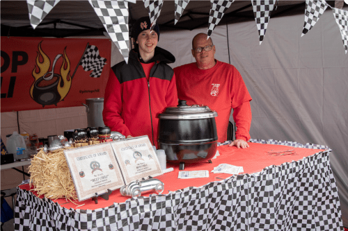 Two men stand behind a table at a chili booth, featuring a black pot and checkered decorations.