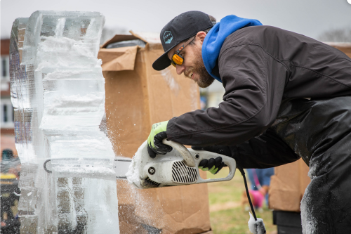 A person in sunglasses and a cap carves an ice block with a chainsaw, focused on their work outdoors.