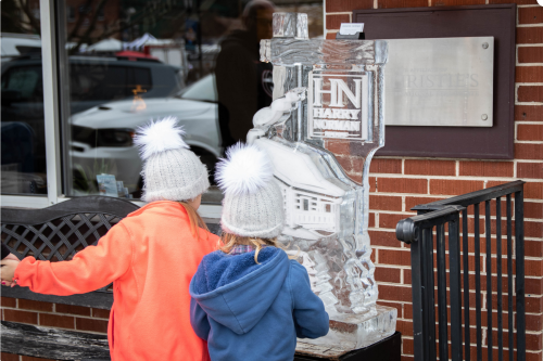 Two children in winter hats admire an ice sculpture outside a brick building.