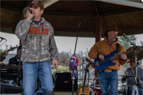 A man sings into a microphone on stage while another plays bass guitar, with instruments and a festive atmosphere around them.