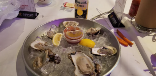 A platter of fresh oysters on ice, accompanied by lemon wedges, cocktail sauce, and a bottle of Corona beer.