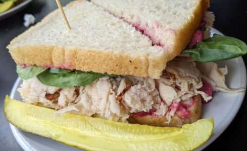 A sandwich with shredded chicken, spinach, and pink spread, served with a pickle on the side.