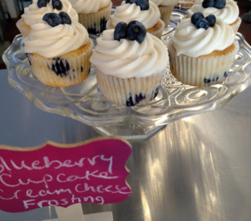 A display of blueberry cupcakes with cream cheese frosting, topped with blueberries, on a glass cake stand.