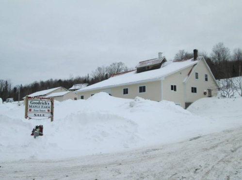 A snowy landscape featuring Goodrich's Maple Farm with a "For Sale" sign in front of a large white building.