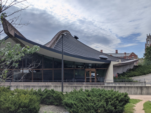 A modern building with a curved roof and large glass windows, surrounded by greenery under a cloudy sky.