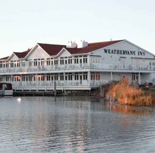 A white inn with a red roof beside a calm lake, surrounded by autumn foliage and reflecting in the water.