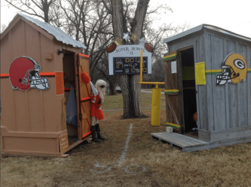 Two themed outhouses representing NFL teams, with a scoreboard showing a Super Bowl game. A person in a cheerleader outfit stands between them.