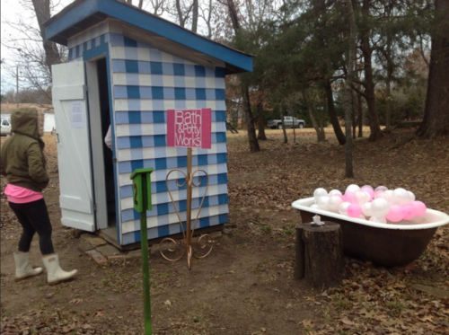 A blue and white checkered shed with a sign reading "Bath & Body Works" next to a bathtub filled with pink balloons.