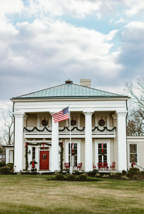 A festive two-story house with a red door, decorated for the holidays, featuring columns and an American flag.