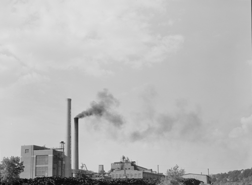 Black and white image of a factory with a tall smokestack emitting smoke against a cloudy sky.