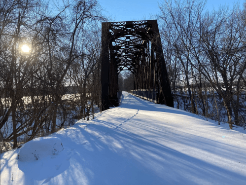 A snow-covered landscape featuring a metal bridge over a frozen river, with sunlight shining through trees.