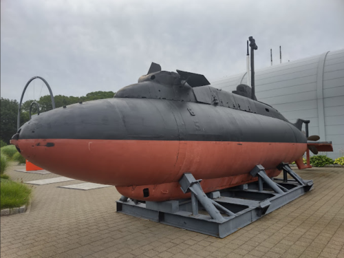 A black and red submarine displayed on a concrete platform, surrounded by greenery and a modern building in the background.