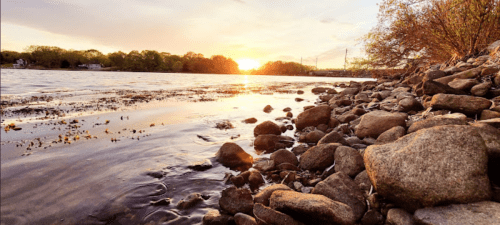 Sunset over a calm river, with a rocky shoreline and trees in the background, reflecting warm colors in the water.