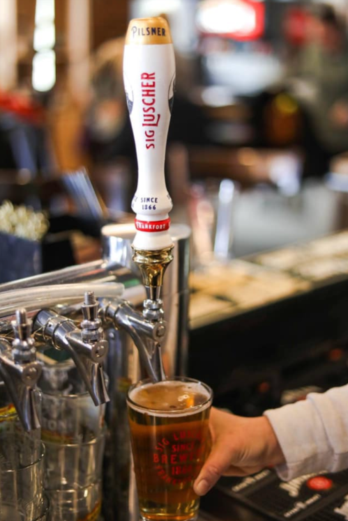 A hand holds a glass of beer under a pilsner tap in a bar setting.