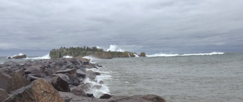 A rocky shoreline with waves crashing against the rocks, leading to a distant island under a cloudy sky.