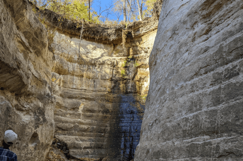 A person stands in a canyon with steep, layered rock walls and a small waterfall cascading down.