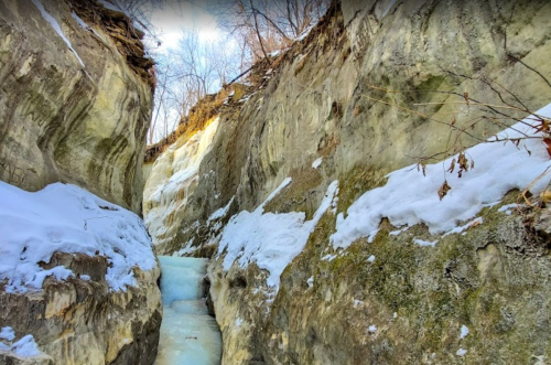 A narrow, icy canyon with snow-covered walls and a frozen stream running through it, surrounded by bare trees.