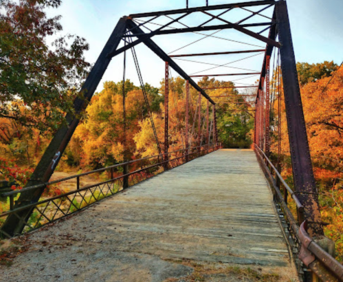 A rustic metal bridge spans a path surrounded by vibrant autumn foliage in warm colors.