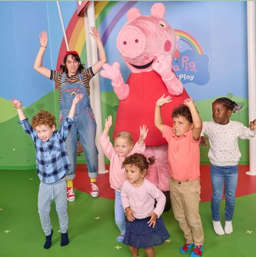 A group of children and an adult pose joyfully with a large Peppa Pig character in a colorful play area.