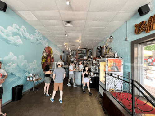 A vibrant café interior with a mural, people ordering, and a cozy seating area. Bright and inviting atmosphere.