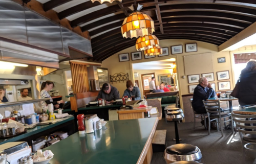 A cozy diner interior with a green counter, patrons seated, and staff preparing food in a warm, inviting atmosphere.