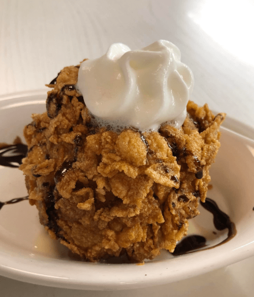 A dessert ball coated in crispy cereal, topped with whipped cream and drizzled with chocolate sauce.