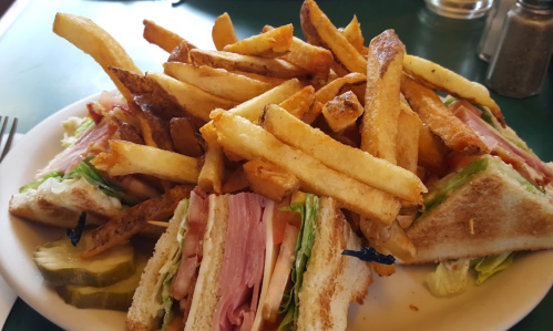 A plate of sandwiches with layers of ham, lettuce, and pickles, topped with a generous serving of golden French fries.