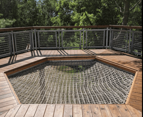 A wooden deck with a hexagonal netted area surrounded by a railing, set in a green, wooded environment.