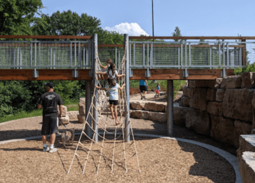 Children play on a rope climbing structure in a park, with a wooden bridge and rocky landscape in the background.