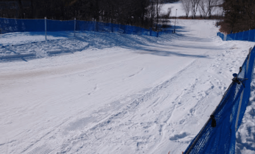 A snowy slope with a blue fence lining the sides, surrounded by trees and a clear sky.