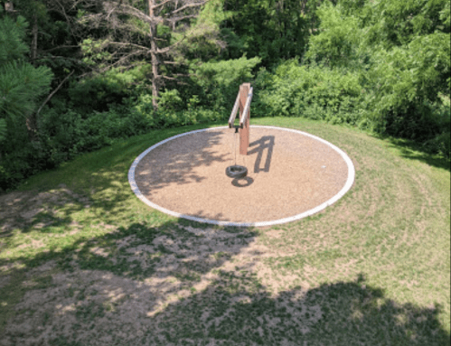 Aerial view of a circular playground area with a tire swing surrounded by grass and trees.