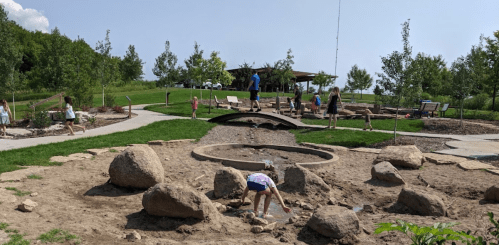 Children play in a park with a sandy area, rocks, and greenery, while adults relax in the background.