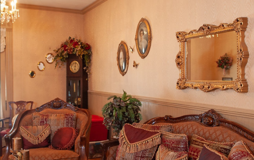 A vintage living room with ornate furniture, decorative mirrors, and floral arrangements on the walls.