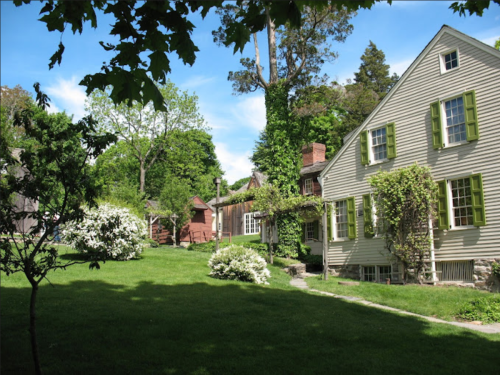 A scenic view of a green lawn with blooming flowers and historic houses under a blue sky with fluffy clouds.