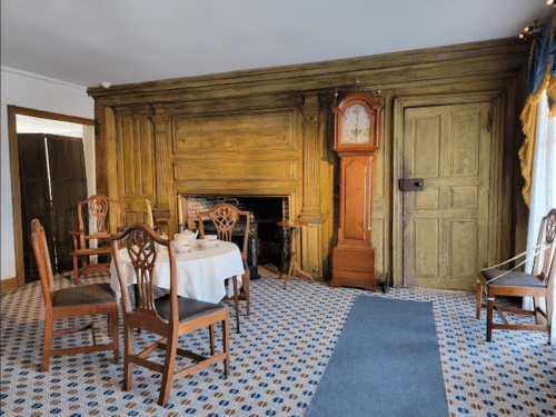 A cozy room featuring a wooden fireplace, a grandfather clock, and a small dining table with chairs.